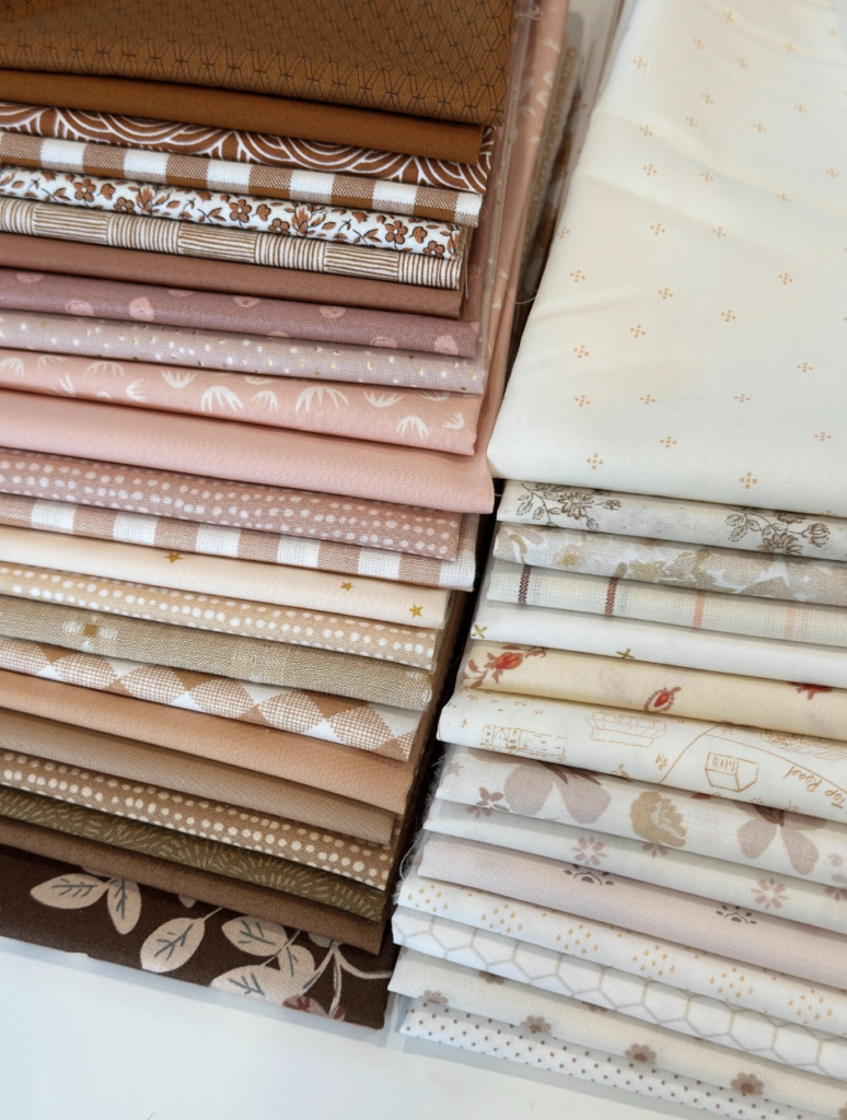 A beautifully arranged stack of quilting fabrics in rich browns, blush pinks, warm neutrals, and creamy low-volume prints. The bundles include a mix of solids, florals, gingham, polka dots, and delicate motifs—perfect for creating a soft and cozy Front Porch Quilt.